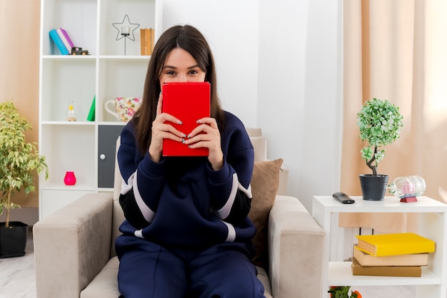 Jeune jolie femme de race blanche assise sur un fauteuil dans un salon conçu tenant un livre et à la recherche de derrière