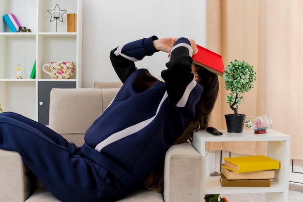 Jeune jolie femme de race blanche assise sur un fauteuil dans un salon conçu toucher la tête avec livre