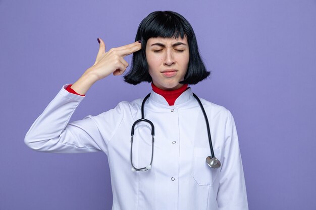 Une jeune jolie femme de race blanche mécontente en uniforme de médecin avec stéthoscope met la main sur son pistolet gestuel de la tempe