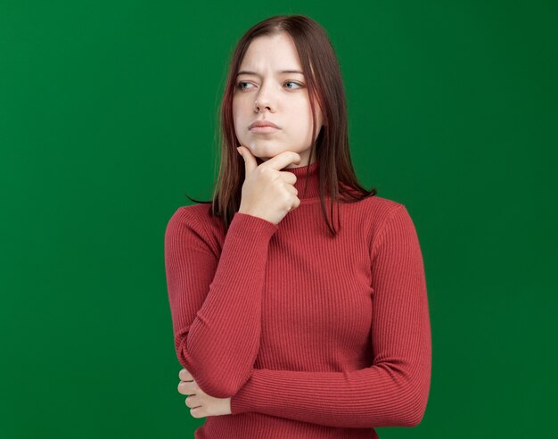 Jeune jolie femme réfléchie gardant la main sur le menton en regardant le côté isolé sur un mur vert avec espace pour copie