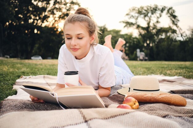 Jeune jolie femme souriante allongée sur un plaid lisant rêveusement un livre sur un pique-nique dans le magnifique parc de la ville