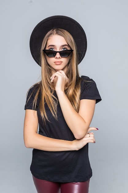 Jeune jolie femme souriante dans des lunettes de soleil brillantes habillées en chapeau noir, t-shirt noir et pantalon sombre isolé sur fond gris poing sous le menton