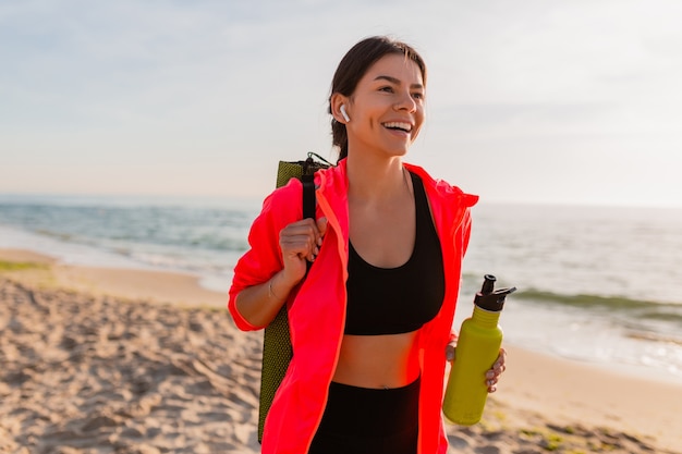 Jeune jolie femme souriante, faire du sport au lever du soleil du matin sur la plage de la mer tenant un tapis de yoga et une bouteille d'eau, mode de vie sain, écouter de la musique sur les écouteurs, porter une veste coupe-vent rose