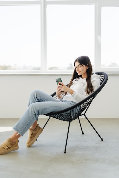 Jeune jolie femme avec un téléphone portable assis dans une chaise papasan près de la fenêtre à la maison