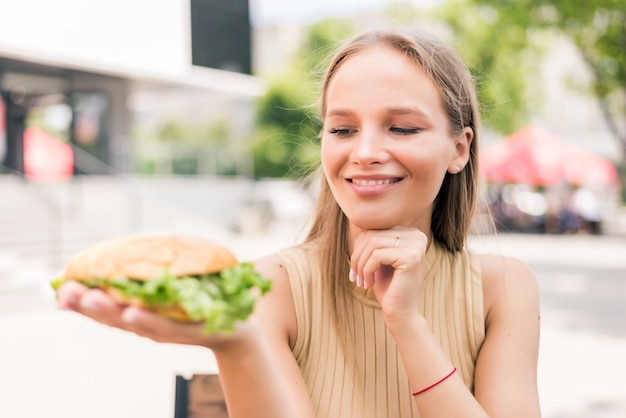 Jeune jolie femme tenant un hamburger assis à l'extérieur