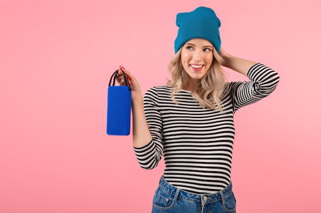 Jeune jolie femme tenant un haut-parleur sans fil écoutant de la musique portant une chemise rayée et un chapeau bleu souriant bonne humeur positive posant sur fond rose