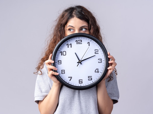 Jeune jolie femme tenant une horloge regardant de côté par derrière elle isolée sur un mur blanc