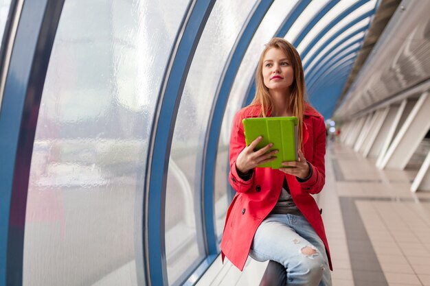 Jeune Jolie Femme En Tenue Décontractée Tenant Un Ordinateur Portable Tablette Dans Un Bâtiment Urbain, Portant Des Jeans, Veste Trench Rose, à La Fenêtre Avec Vue Sur La Ville, L'éducation En Ligne