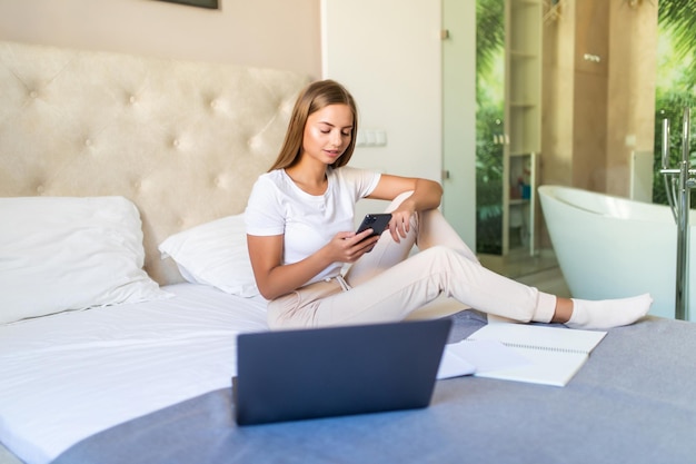 Jeune jolie femme utilise un téléphone et un ordinateur portable assis sur le lit