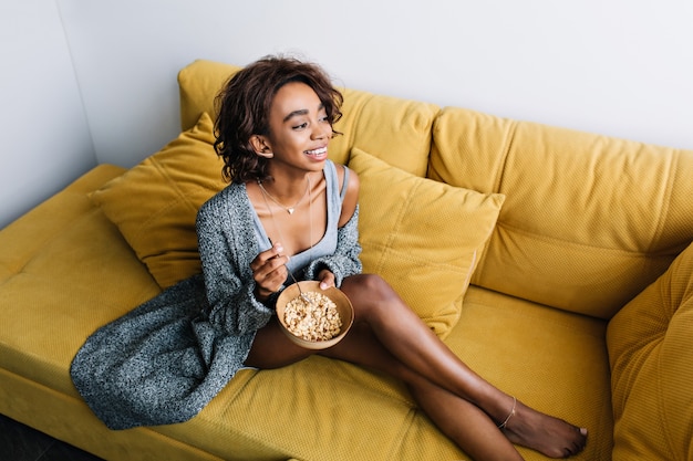 Photo gratuite jeune jolie fille africaine appréciant un petit-déjeuner sain, mangeant du granola assis sur un canapé jaune, un canapé, une atmosphère matinale à la maison. elle a les cheveux courts et bouclés, porte un cardigan décontracté gris, un short.