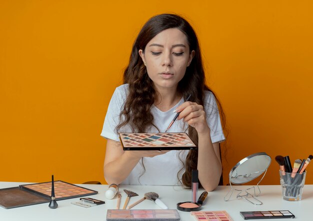 Jeune jolie fille assise à la table de maquillage avec des outils de maquillage tenant la palette de fard à paupières et le pinceau et les regardant isolés sur fond orange