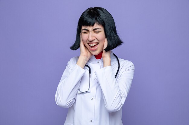 Jeune jolie fille caucasienne agacée en uniforme de médecin avec stéthoscope couvrant ses oreilles avec les mains