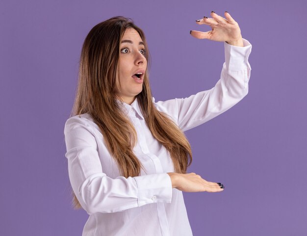 Une jeune jolie fille caucasienne choquée garde les mains ouvertes en faisant semblant de tenir quelque chose d'isolé sur un mur violet avec un espace de copie