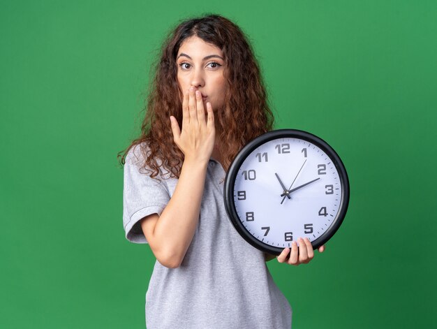 Jeune jolie fille caucasienne concernée tenant une horloge en gardant la main sur la bouche isolée sur un mur vert avec espace pour copie