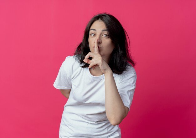 Jeune jolie fille caucasienne gardant la main derrière le dos et faisant le geste de silence à la caméra isolée sur fond cramoisi avec espace de copie