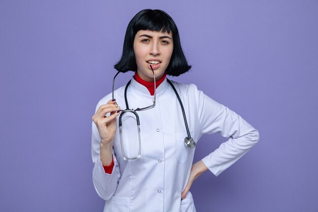 Jeune jolie fille caucasienne ignorante en uniforme de médecin avec stéthoscope tenant des lunettes optiques et regardant à l'avant