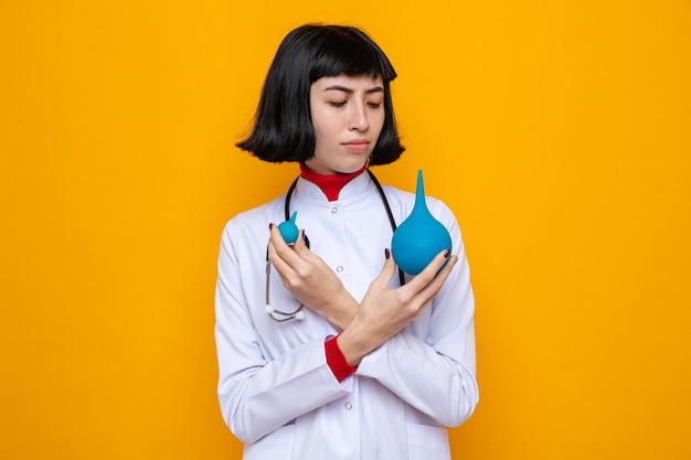 Jeune jolie fille caucasienne ignorante en uniforme de médecin avec stéthoscope tenant et regardant des lavements