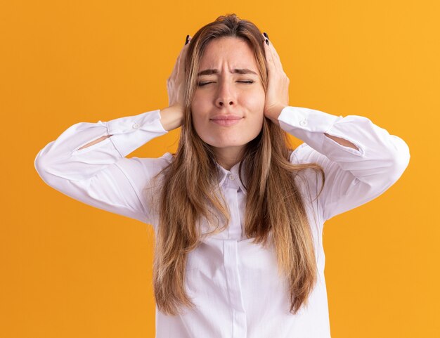 Une jeune jolie fille caucasienne mécontente ferme les oreilles avec les mains isolées sur un mur orange avec espace de copie