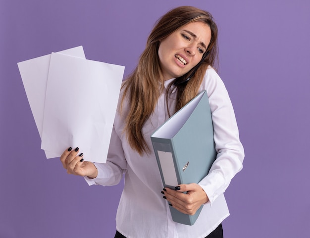 Une jeune jolie fille caucasienne mécontente tient des feuilles de papier et un dossier parlant au téléphone isolé sur un mur violet avec espace de copie