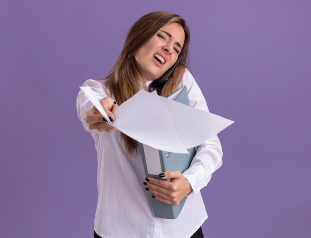 Une jeune jolie fille caucasienne mécontente tient des feuilles de papier en gardant le dossier et en parlant au téléphone