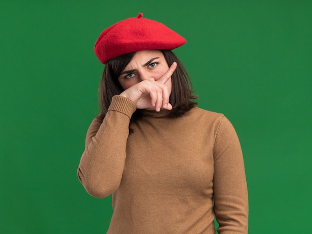 Photo gratuite une jeune et jolie fille caucasienne sérieuse avec un chapeau de béret met la main sur le nez isolé sur un mur vert avec espace de copie