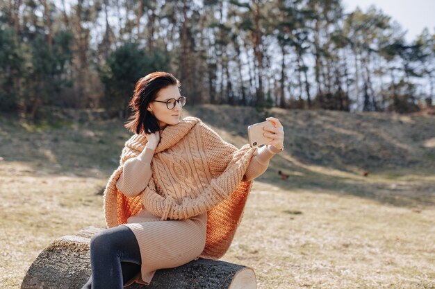 Jeune jolie fille élégante sur la nature sur le mur de la forêt avec un téléphone par une journée ensoleillée en prenant des photos d'elle-même. vacances en plein air et dépendance à la technologie.