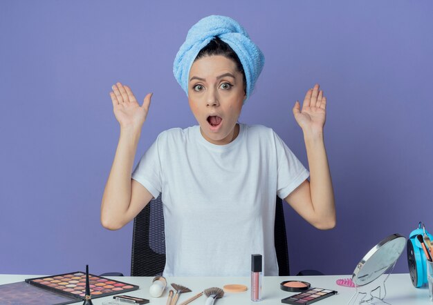 Jeune jolie fille impressionnée assise à la table de maquillage avec des outils de maquillage et avec une serviette de bain sur la tête montrant les mains vides