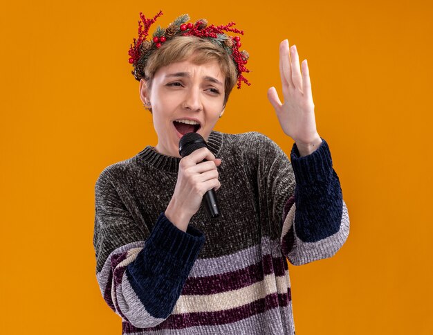 Jeune jolie fille portant une couronne de tête de Noël tenant un microphone près de la bouche en gardant la main dans l'air à côté chant isolé sur mur orange