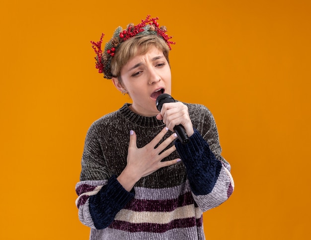 Jeune jolie fille portant une couronne de tête de Noël tenant un microphone regardant vers le bas en gardant la main sur la poitrine chantant isolé sur mur orange avec espace copie