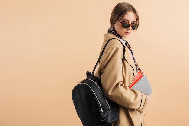 Jeune jolie fille en trench-coat et lunettes de soleil avec sac à dos sur l'épaule tenant des blocs-notes à la main regardant pensivement à huis clos tout en passant du temps sur fond beige