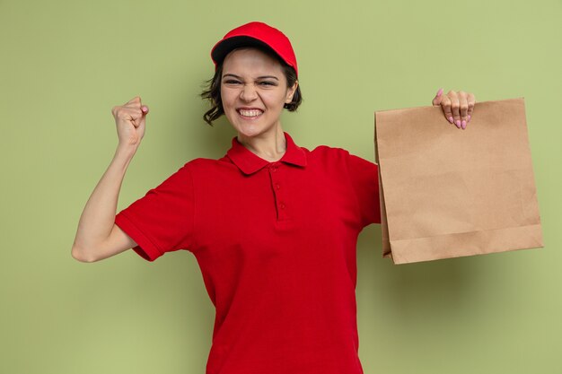 Jeune jolie livreuse excitée tenant des emballages alimentaires en papier et levant le poing