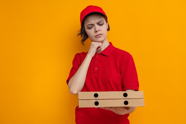Jeune jolie livreuse ignorante tenant et regardant des boîtes à pizza mettant la main sur son menton