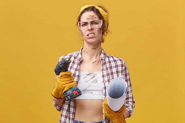Jeune jolie travailleuse portant des lunettes de protection, des gants et une chemise décontractée tenant une machine de forage et un plan ayant un regard dégoûtant fronçant les sourcils avec le refus de réparer sa chambre