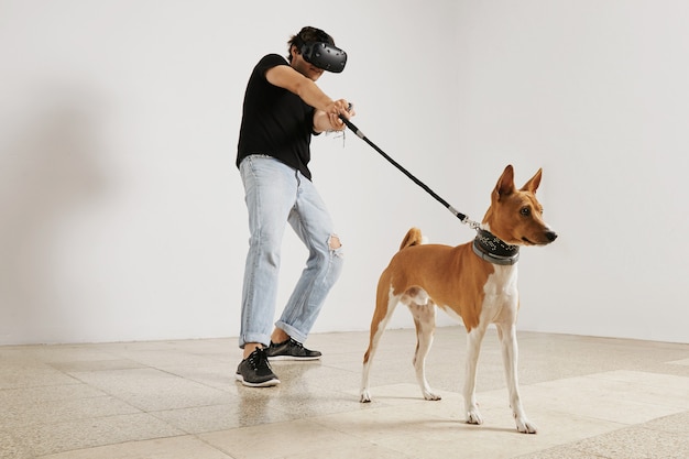 Un jeune joueur en casque VR et t-shirt noir sans étiquette tirant une laisse sur un chien basenji marron et blanc sur un mur blanc.