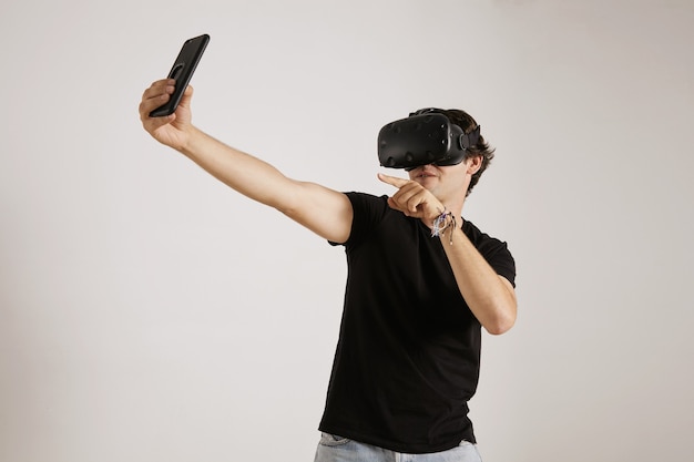 Un jeune joueur en t-shirt noir et casque VR posant pour un selfie sur son smartphone sur un mur blanc