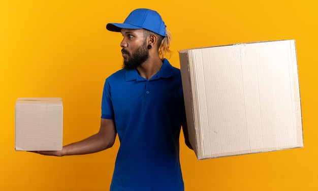 Jeune livreur afro-américain impressionné tenant des boîtes en carton regardant le côté isolé sur un mur orange avec espace de copie