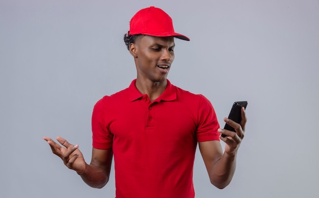 Jeune livreur afro-américain mécontent portant un polo rouge et une casquette en regardant smartphone à la main montrant l'aversion sur blanc isolé