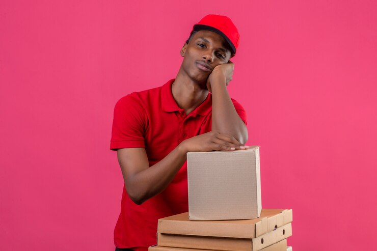 Photo gratuit jeune livreur afro-américain portant un polo rouge et une casquette en attente se pencha son coude sur une boîte en carton sur rose isolé