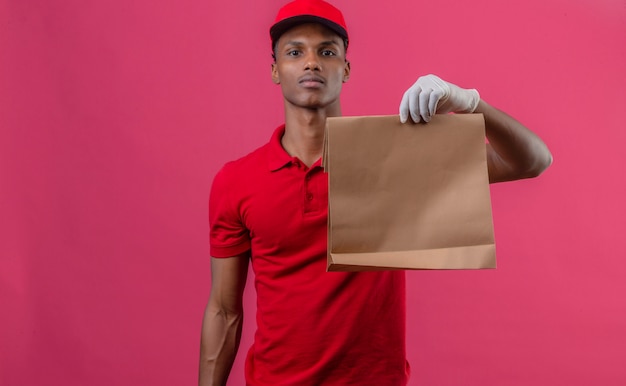 Jeune livreur afro-américain portant un polo rouge et une casquette dans des gants de protection tenant un sac en papier avec des plats à emporter sur rose isolé