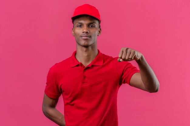 Jeune livreur afro-américain portant un polo rouge et une casquette debout avec le concept du gagnant du poing levé sur rose isolé