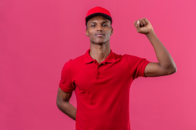 Jeune livreur afro-américain portant un polo rouge et une casquette debout avec le concept du gagnant du poing levé sur rose isolé