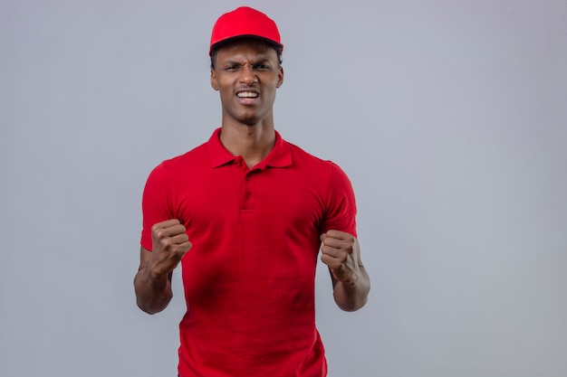 Jeune livreur afro-américain portant un polo rouge et une casquette levant les poings frustrés et furieux sur blanc isolé