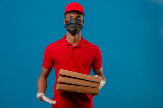 Jeune livreur afro-américain portant un polo rouge et une casquette en masque de protection et des gants debout avec une pile de boîtes à pizza sur bleu isolé