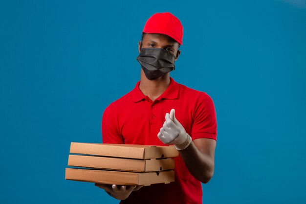 Jeune livreur afro-américain portant un polo rouge et une casquette en masque de protection et des gants debout avec une pile de boîtes à pizza faisant un geste d'argent sur bleu isolé