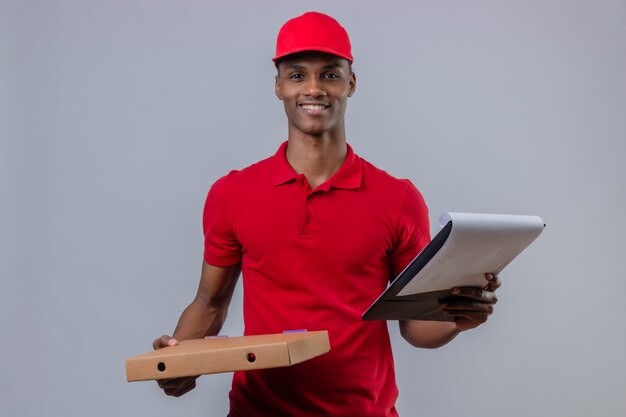 Jeune livreur afro-américain portant un polo rouge et une casquette tenant une boîte à pizza et un presse-papiers avec sourire sur le visage sur blanc isolé