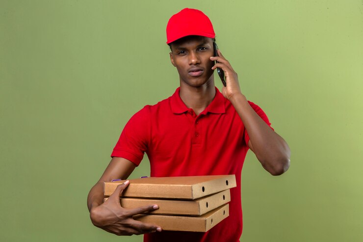 Photo gratuit jeune livreur afro-américain portant un polo rouge et un chapeau portant une pile de boîtes à pizza tout en parlant par smartphone sur vert isolé