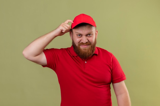 Jeune livreur barbu en uniforme rouge et cap à la confusion et incertain se gratter la tête