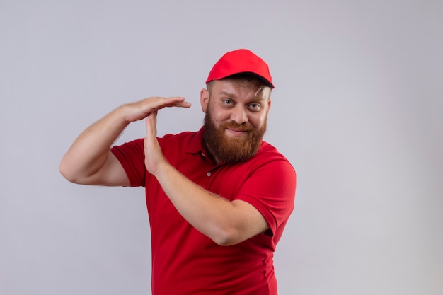 Jeune livreur barbu en uniforme rouge et cap à la fatigue et surmené faisant le geste de temps avec les mains