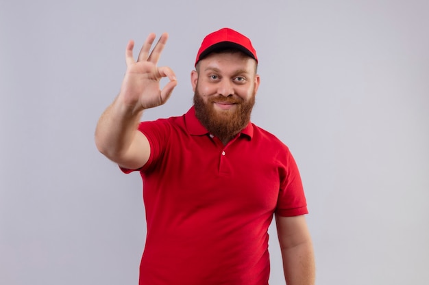 Jeune livreur barbu en uniforme rouge et chapeau souriant positif et heureux de faire signe ok