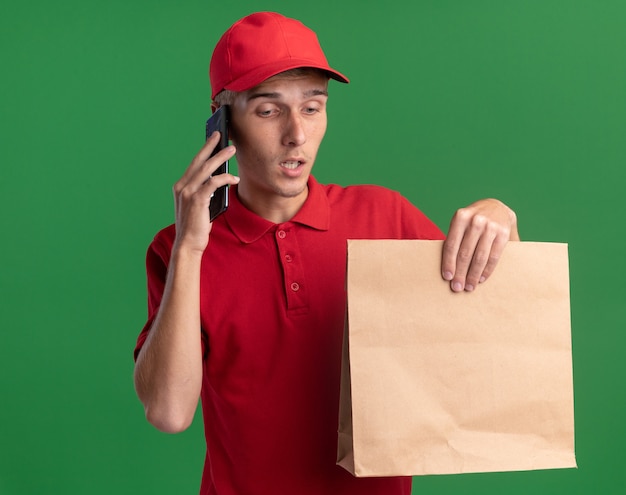 Photo gratuite un jeune livreur blond déçu tient un paquet de papier et parle au téléphone